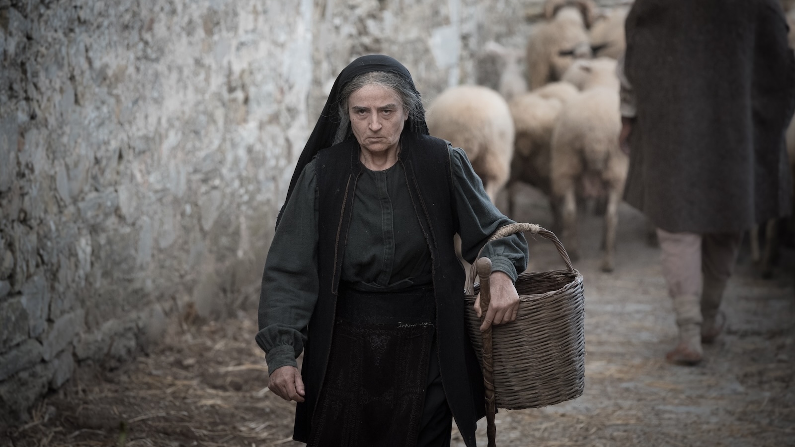 An angry looking older woman carries a basket and wears a scarf at the back of her head as she walks down a road in a city with sheep behind her