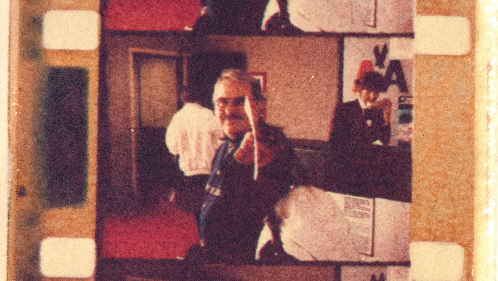 Old color film reel image of a man with mustache and glasses holding a piece of paper towards the camera