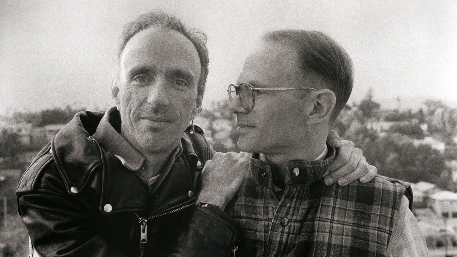 Two men, affectionate with one another, pose for the camera, one looks at us, the other, wearing glasses, looks at him.