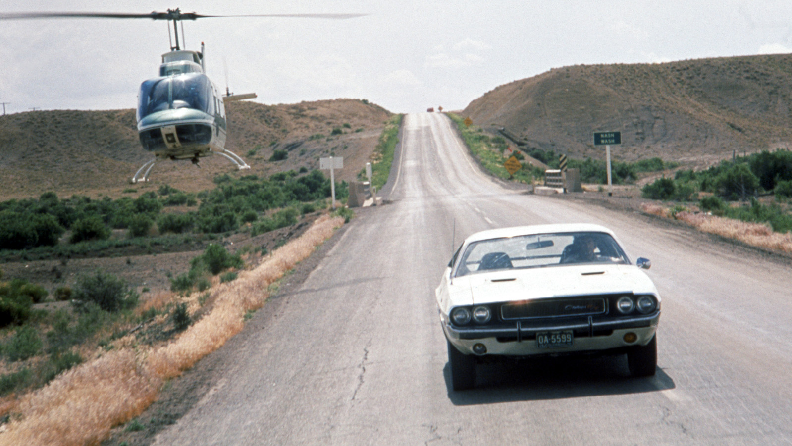 A lone car on a highway, a white Dodge Challenger, pursued by a helicopter.