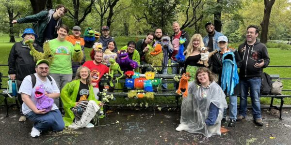 A group of eighteen smiling people holding puppets sit stand and sit by a bench in front of a park. In front of them on the pavement there are colorful stencil drawings of Muppets.
