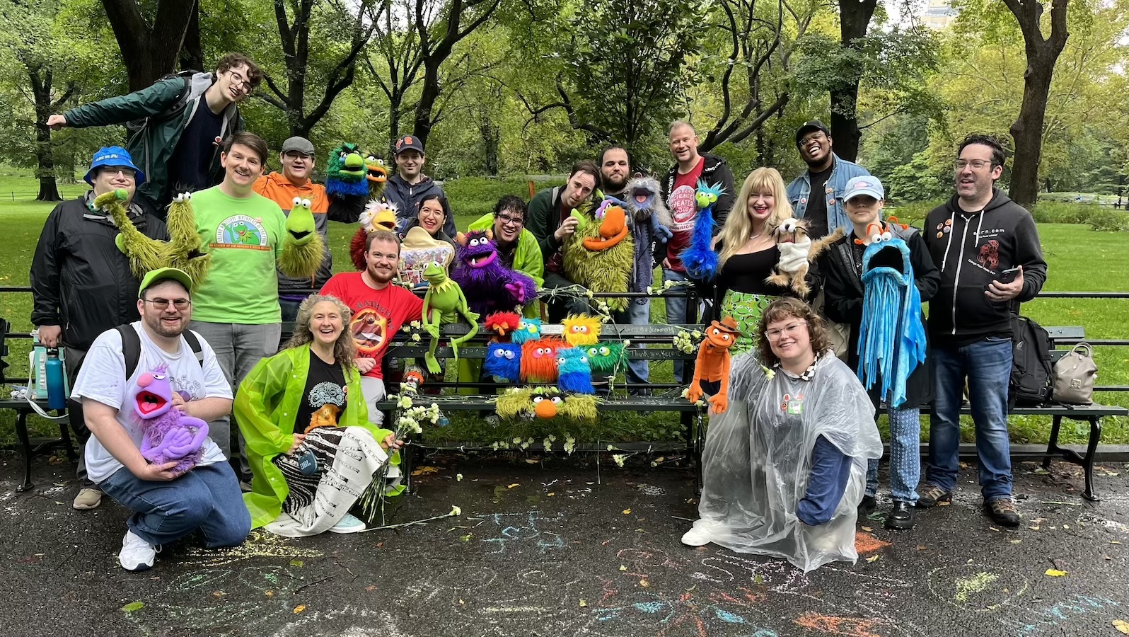 A group of eighteen smiling people holding puppets sit stand and sit by a bench in front of a park. In front of them on the pavement there are colorful stencil drawings of Muppets.