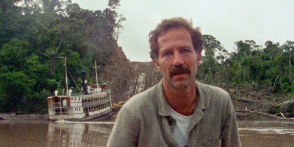 A man with a mustache stands before a jungle river, as a large riverboat sits in the water behind him