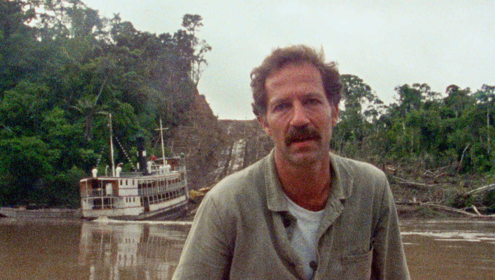 A man with a mustache stands before a jungle river, as a large riverboat sits in the water behind him