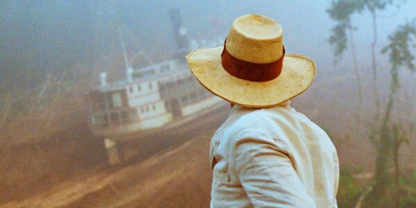 A figure in a pale yellow panama hat and white jacket turns away to look at a large riverboat scaling down the side of a mountain