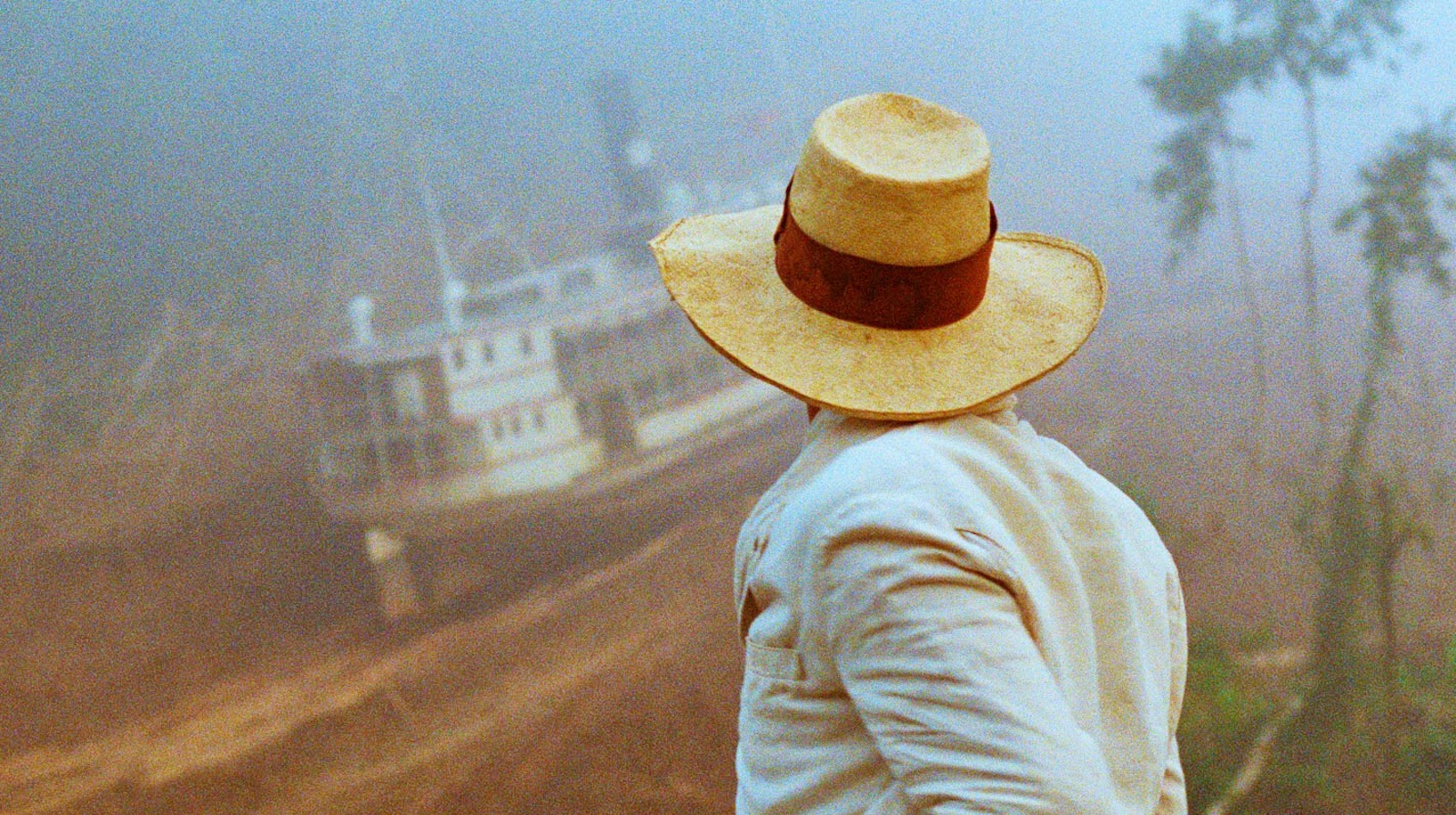 A figure in a pale yellow panama hat and white jacket turns away to look at a large riverboat scaling down the side of a mountain