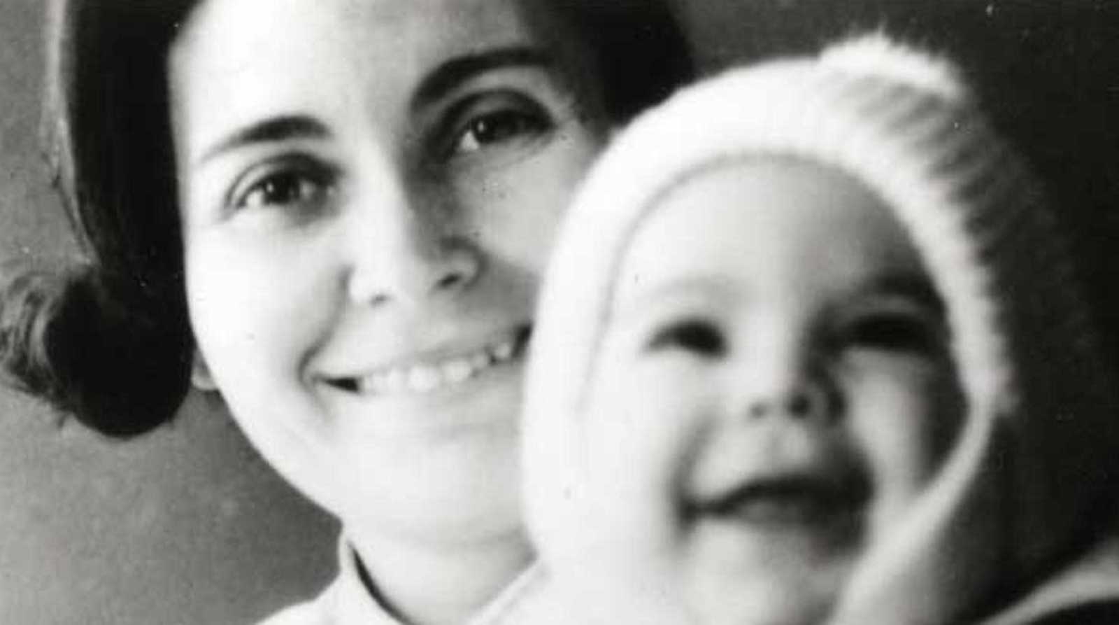 A woman poses with her baby sitting in front of her as they both look in the camera, both smiling