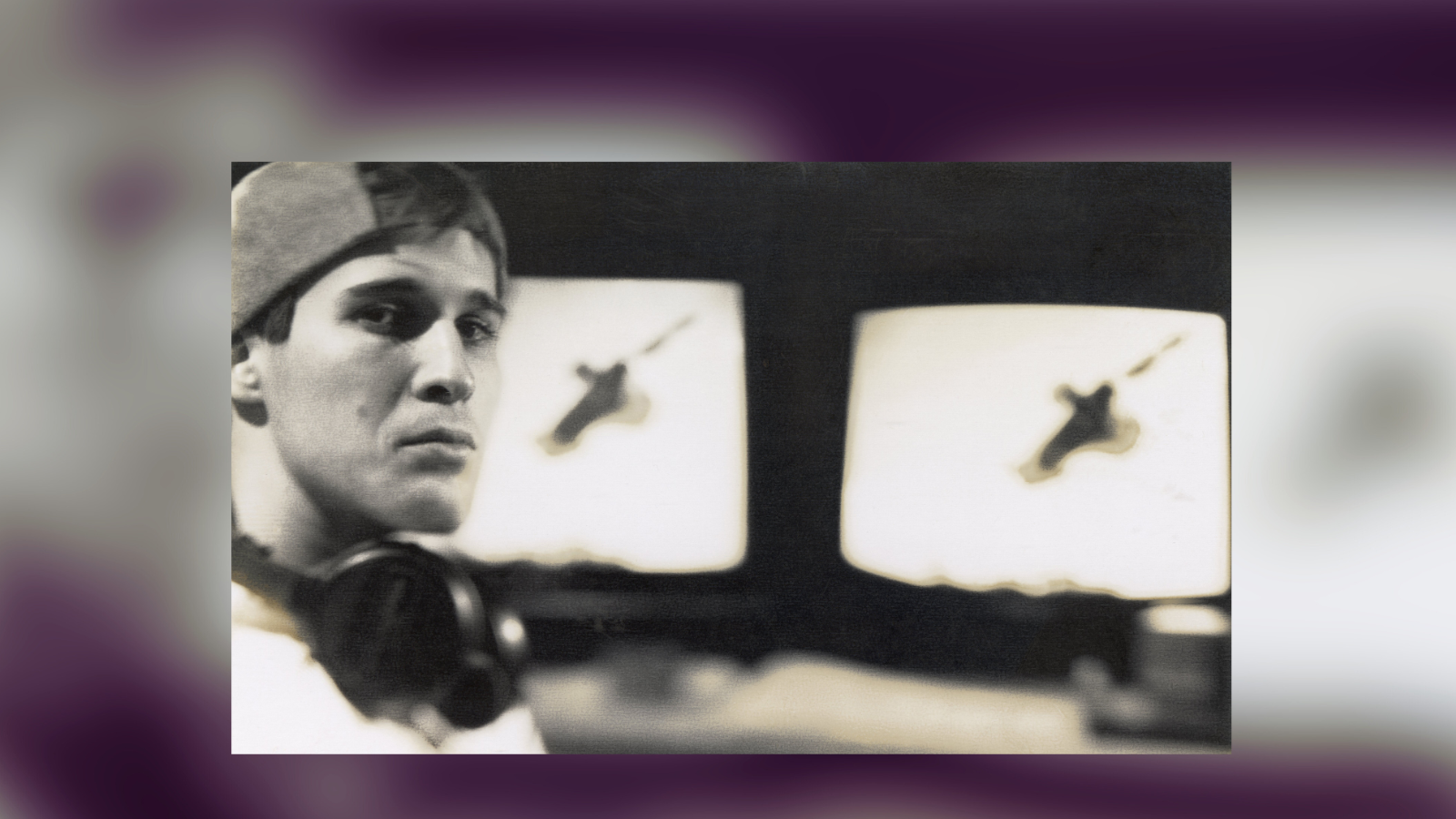 On the left side of the black and white photo, a a person with pale skin wearing a backwards baseball cap on his head and headphones around his next looks at the view while behind him are two large CRT monitors with blurred images of a figure in midair.