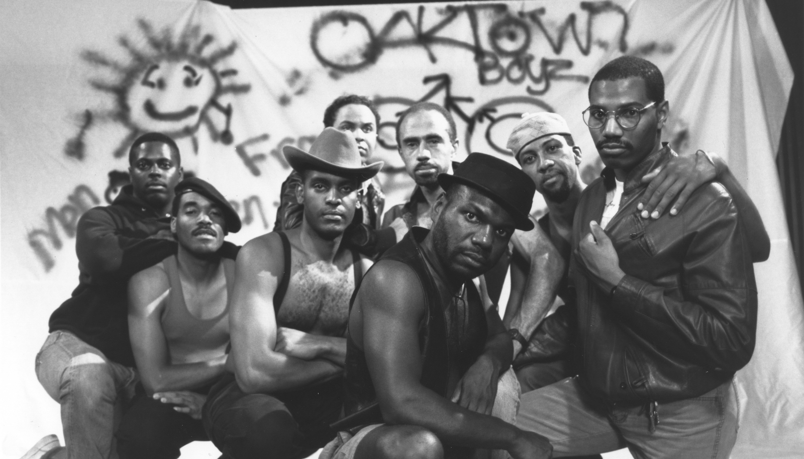 A group of Black men stand together and look in camera in front of a graffiti that says Oaktown Boyz.