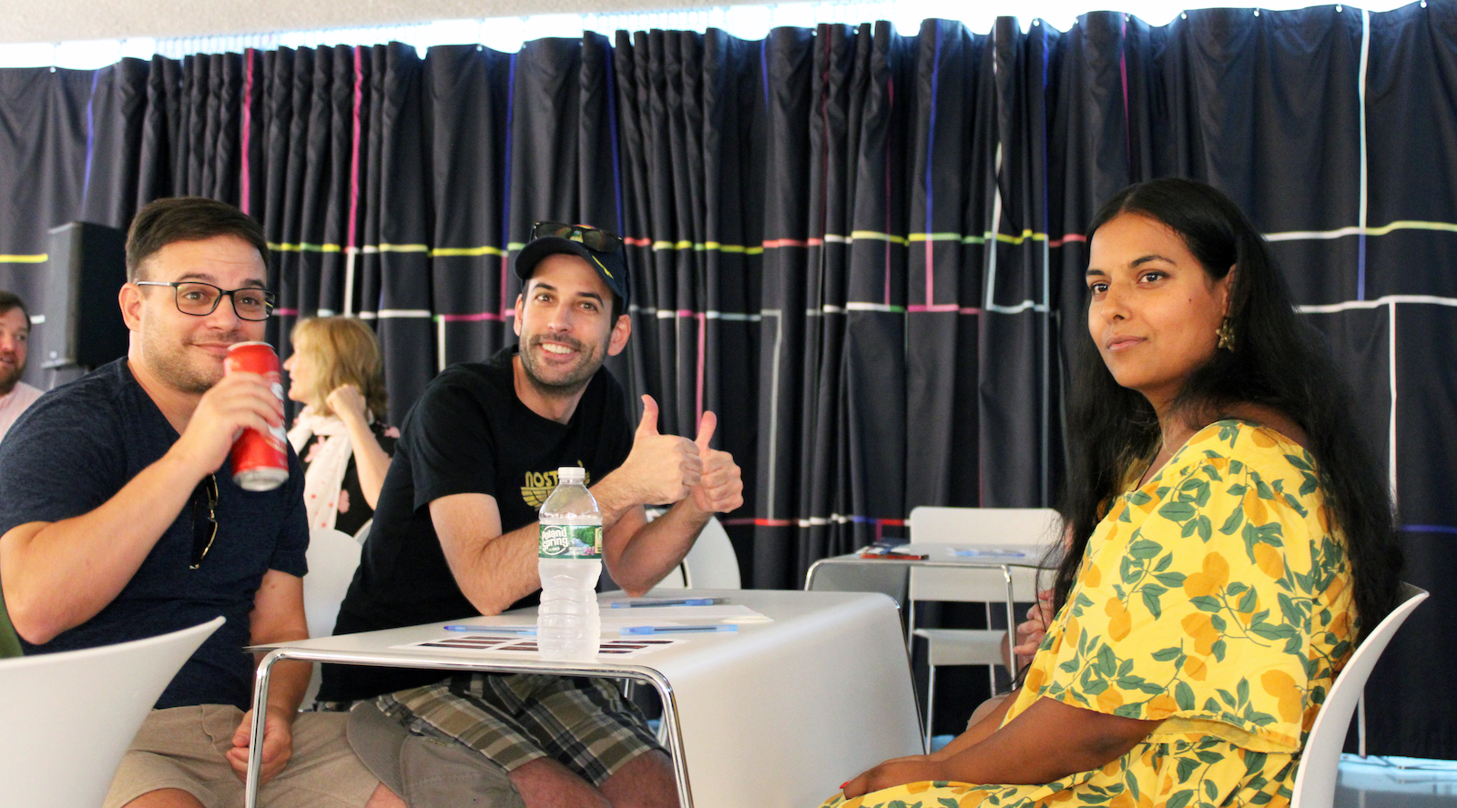 Two men and a woman sit at a white table with a dark curtain behind them. The men on the left wear black T-shirts, the woman on the right wears a bright yellow floral dress. The man in the middle, wearing a ballcap gives the thumbs up.