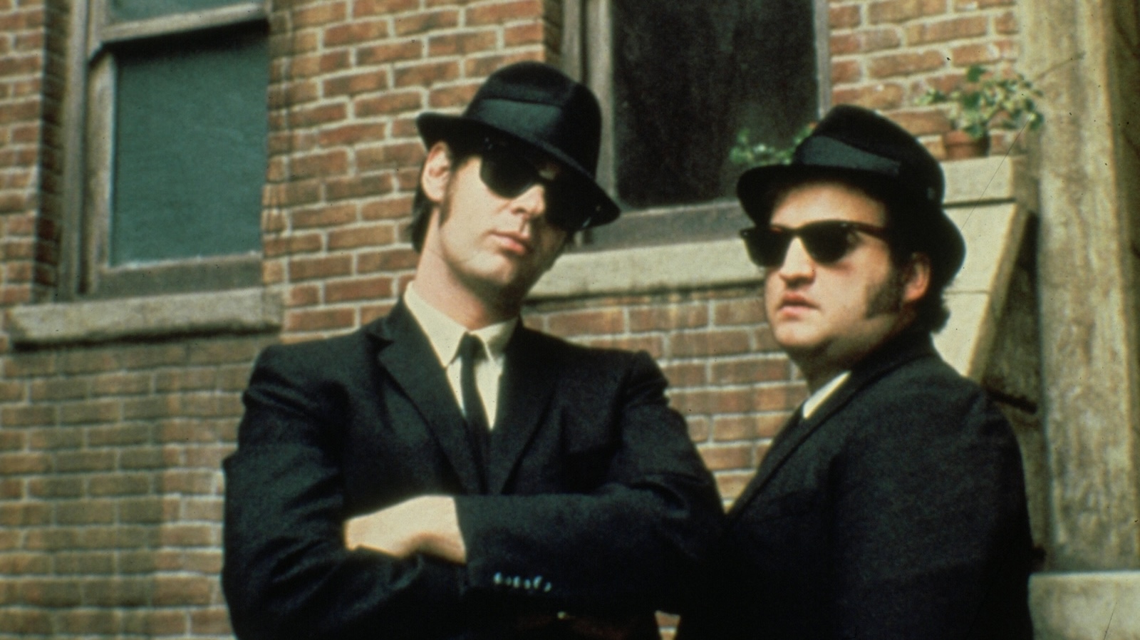 Two men in sunglasses, fedoras, and suits look at us, the one on the left crossing his hands