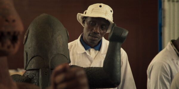 A man in a white hat thoughtfully looks at an African statue in a museum.