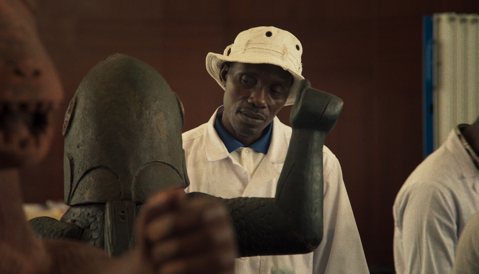 A man in a white hat thoughtfully looks at an African statue in a museum.