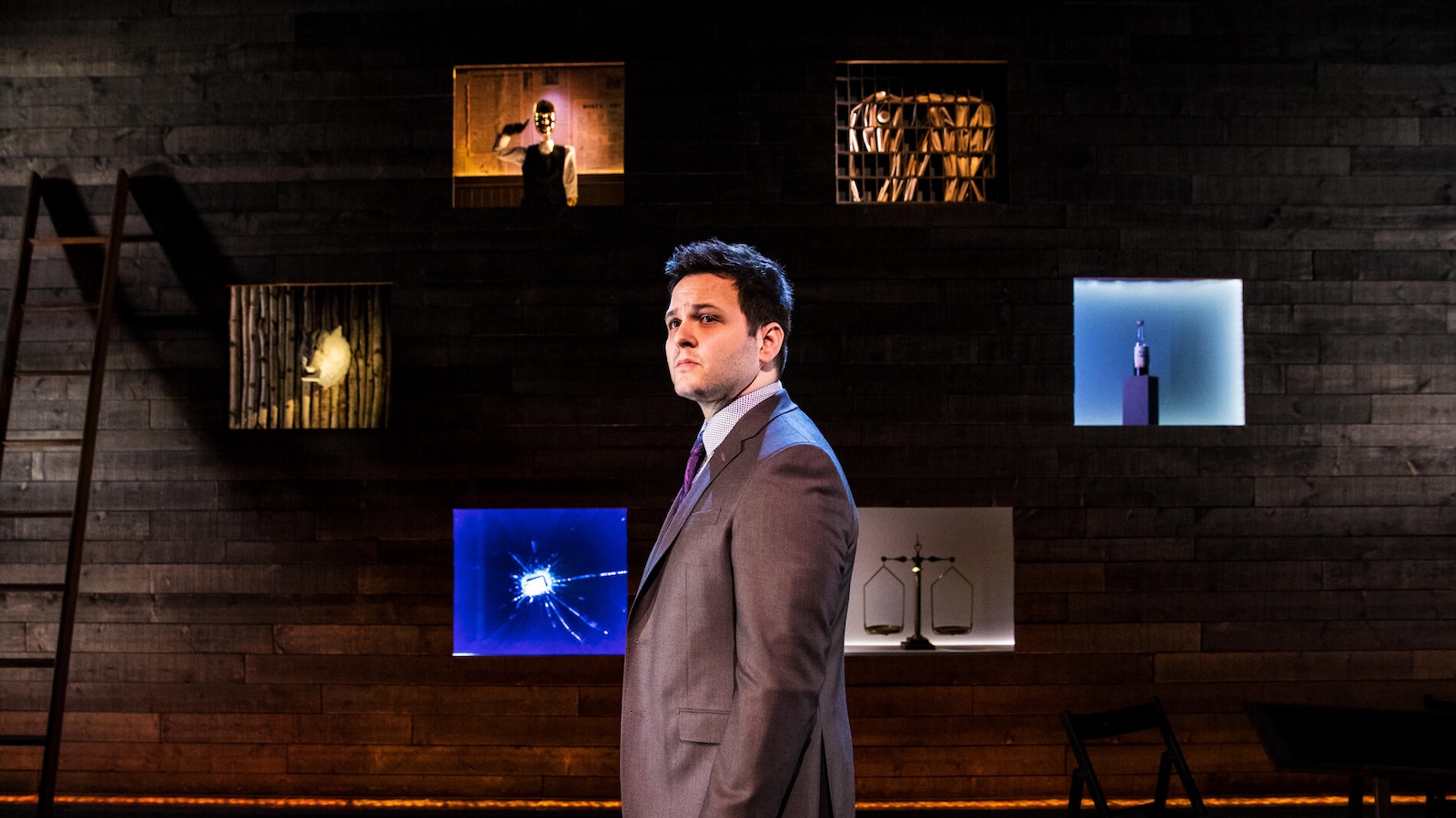 A man in a suit stands in front of six illuminated screens against a dark brown wall.