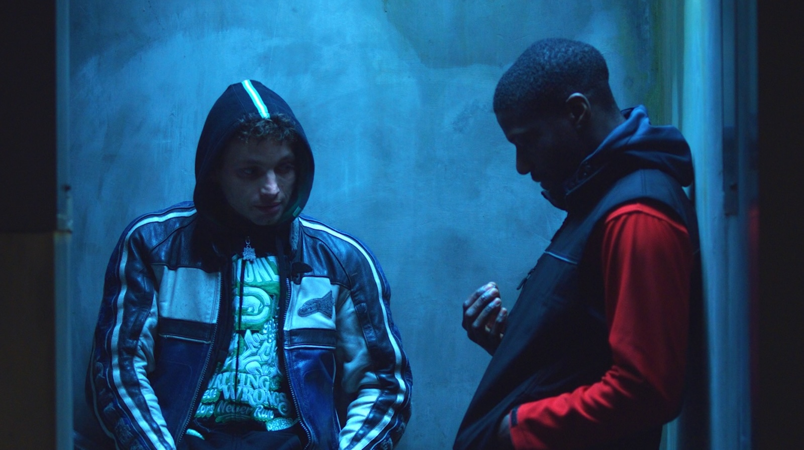Two young men, one in a hooded jacket talk in a blue-tinted nighttime alley.