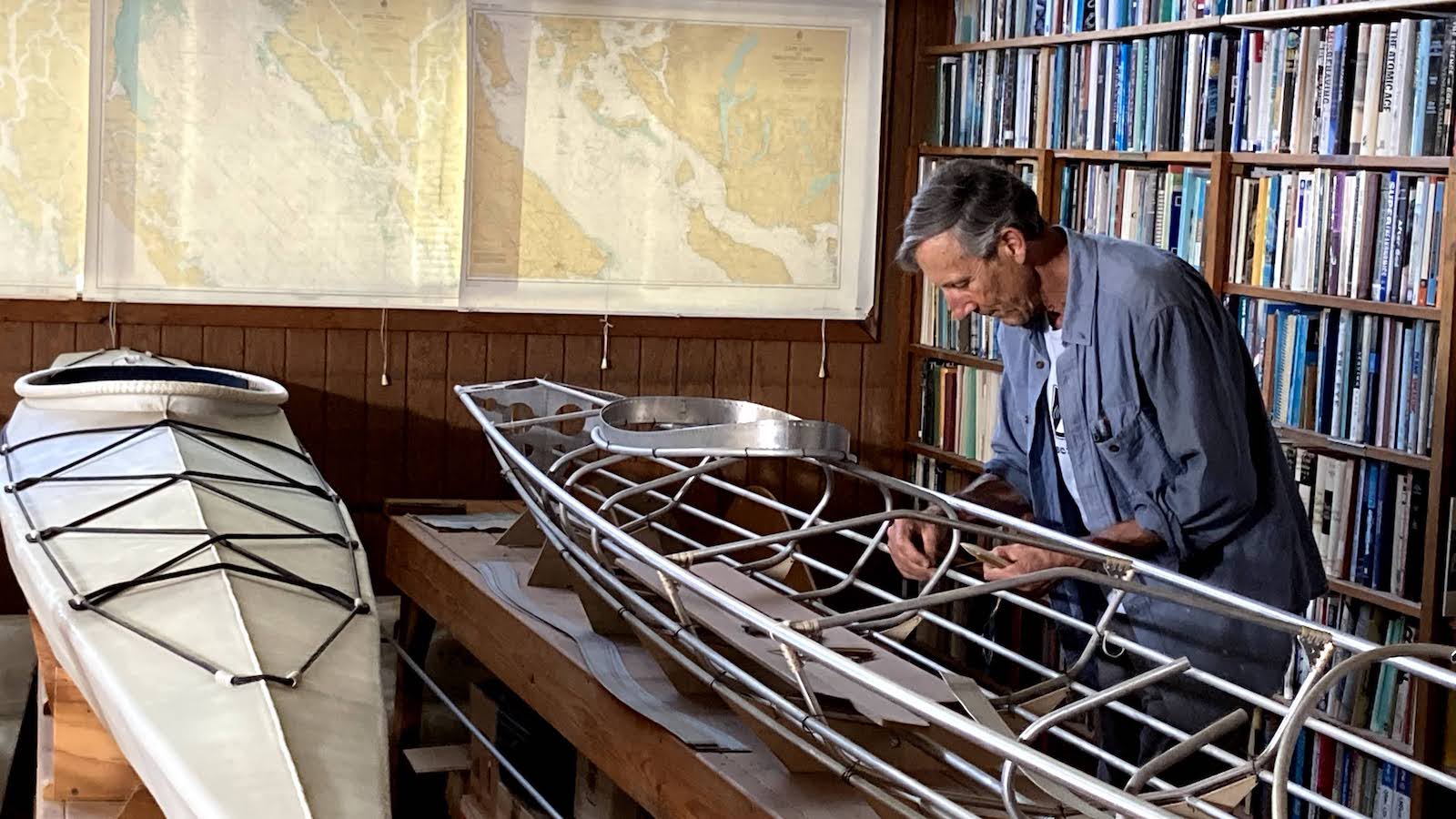 A man in an open blue button down over a white t-shirt stands in a library in front of a bookshelf and hovers over a skeletal metal model of a canoe, which is next to a white canoe on another table