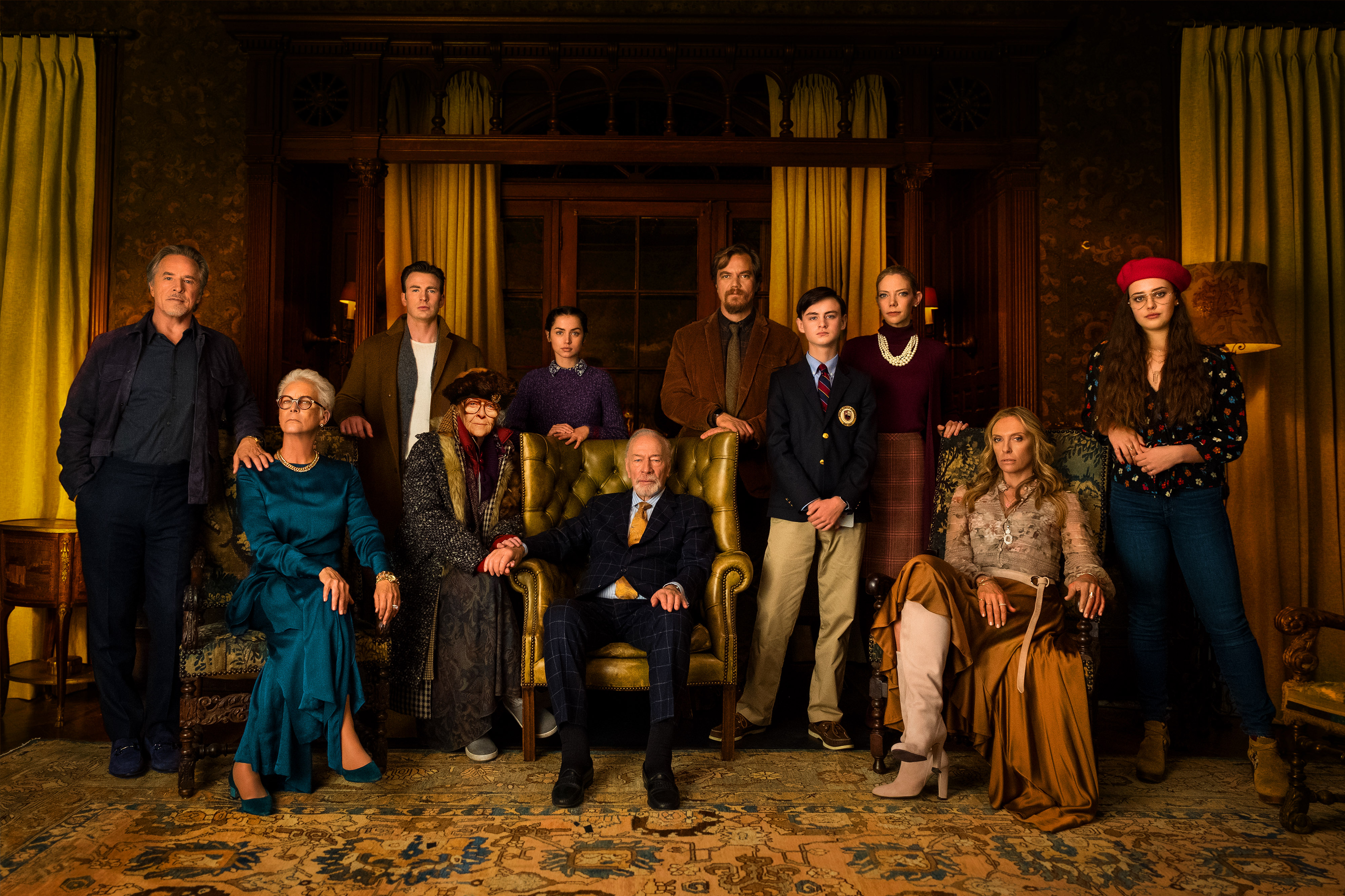 A stern looking family of 12 people sit and stand in a beige and yellow colored library on an ornate rug. In the center is an elderly man in a suit who is clearly the patriarch. All seem to be posing for a picture.