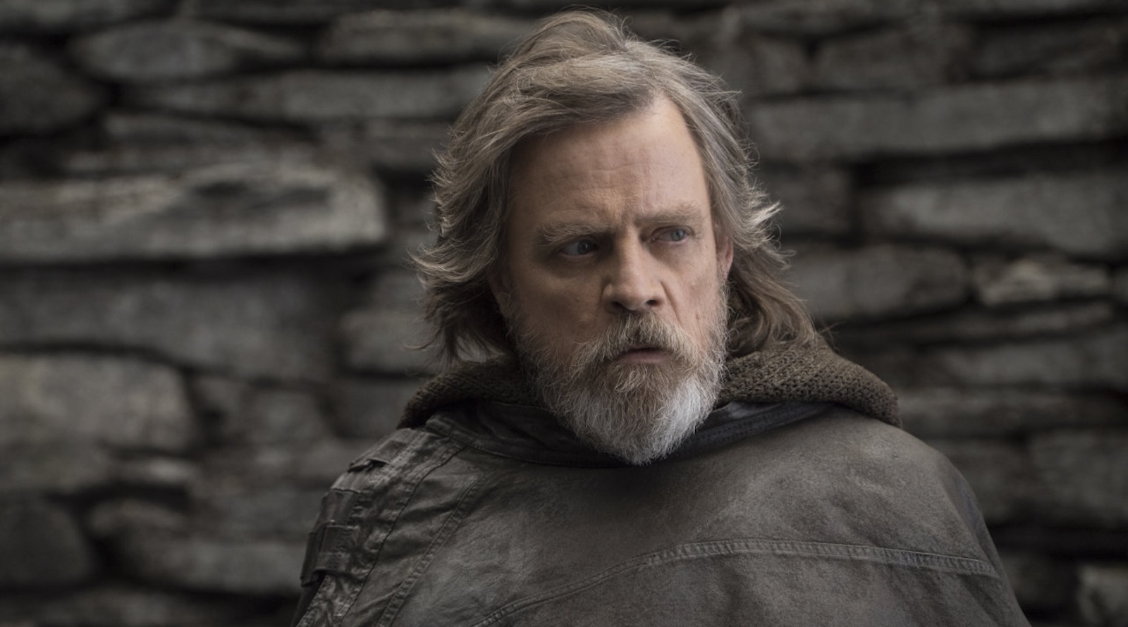 A bearded man in a cloak with a serious expression stands against a stone wall