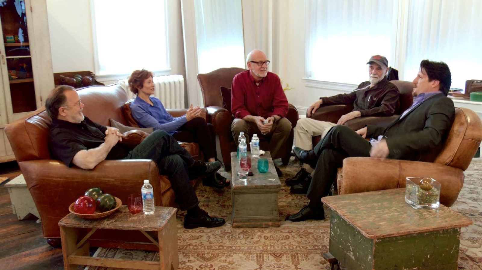 Five people, four men and a woman, sit in reclining chairs around a small table talking in a sunlit room
