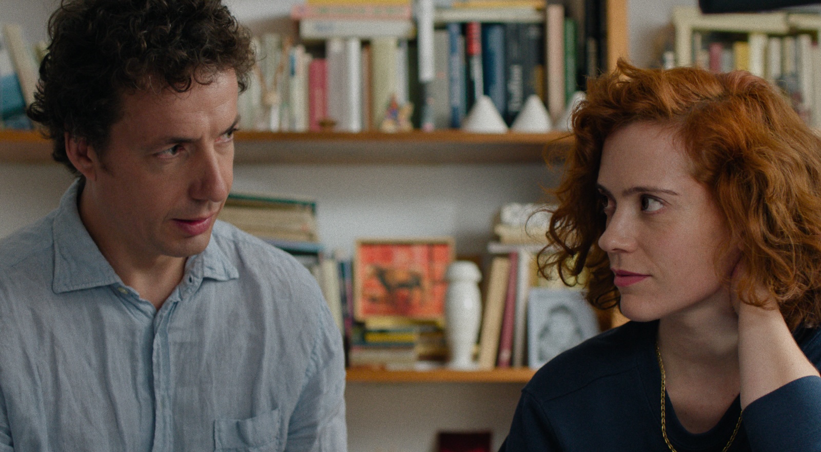 A redheaded woman and a dark haired man sit against a bookshelf backdrop and look tentatively at each other.