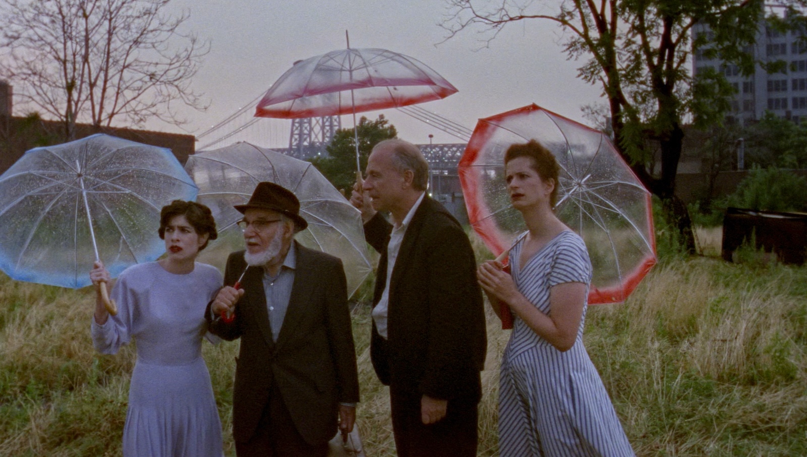 Four people stand in a parched field amidst city buildings holding transparent umbrellas.