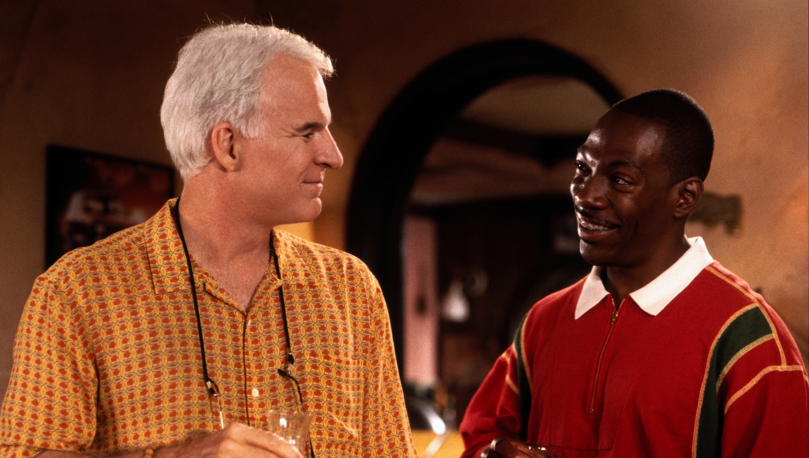 Two men stand and smile at each other in a beige colored room, the man in the left in a yellow shirt and glasses around his neck attached to a string, the one on the right wearing a red shirt with a white collar.