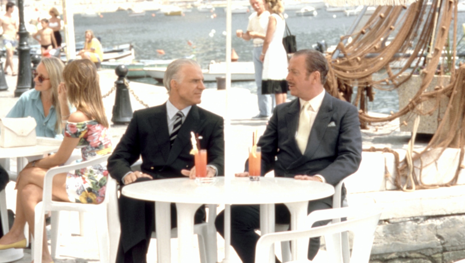 Two fashionably suited men with slick backed hair sit with cocktails at an outdoor restaurant on the docks