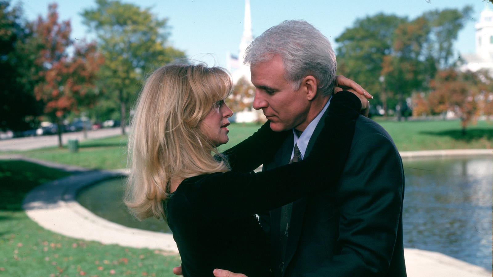A white-haired man in a dark suit and a blonde woman in a black shirt embrace against an outdoor New England backdrop near a fountain and a church spire in the distance