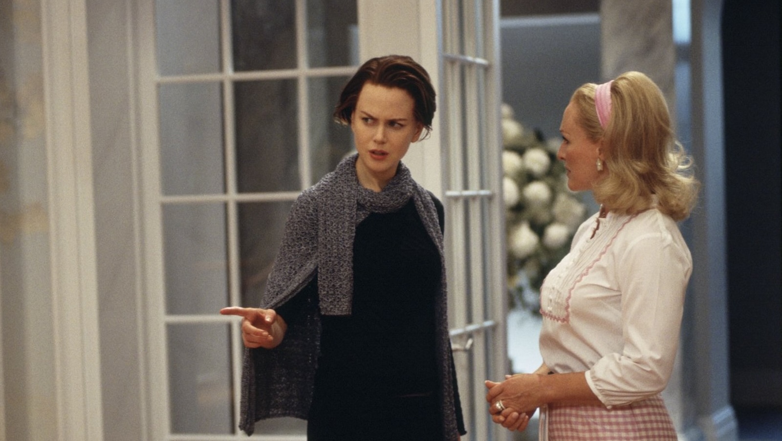 A brunette woman in a black shirt and dark grey sweater gestures suspiciously with her hand while standing next to a brightly dressed blonde woman with a pink hair ribbon, white shirt and gingham skirt