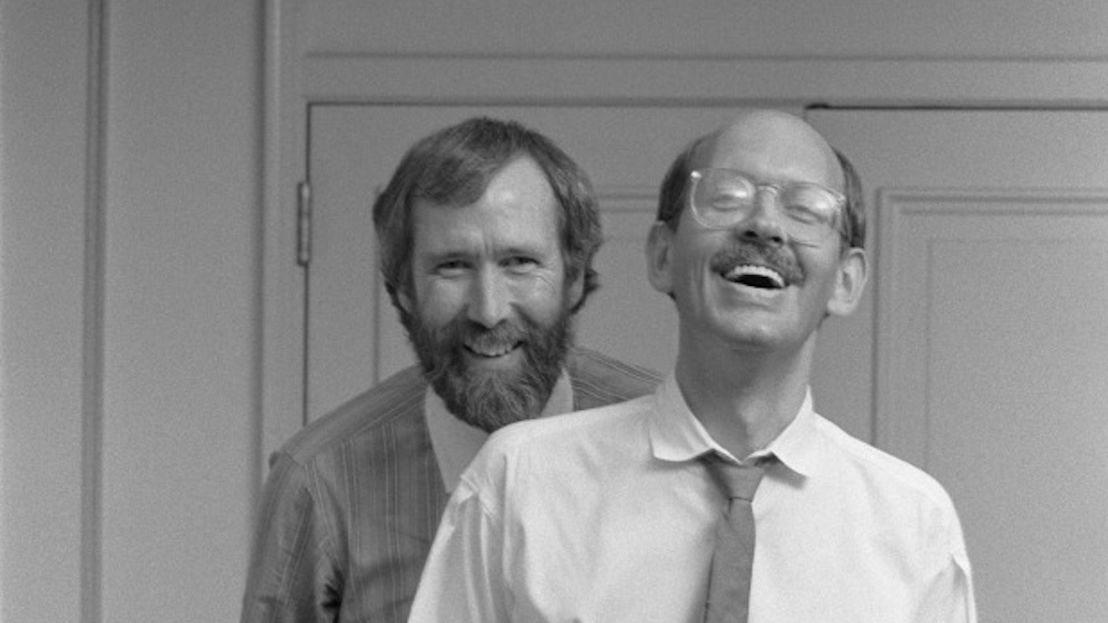Two men—one bearded and one clean-shaven—smile joyously in a black and white photo.