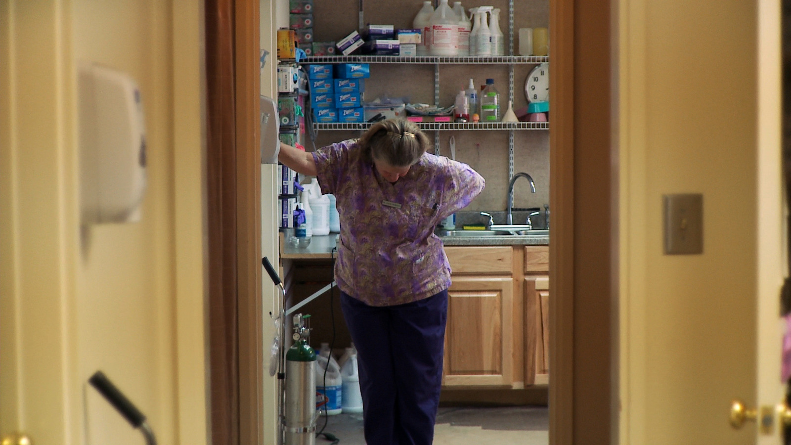 In a hospital back room, an exhausted middle-aged woman in a purple and white shirt puts her hand on a wall and hangs her head in fatigue.