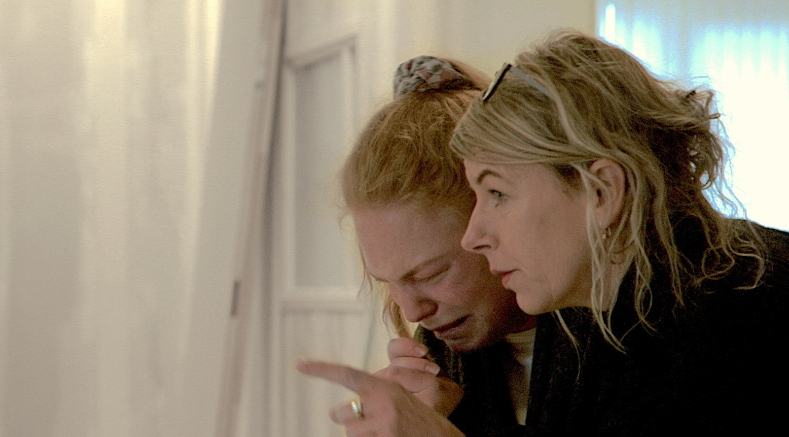 Two women in profile, one grasping her hands in pain and discomfort, the other staring ahead.