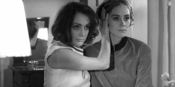 Two women sit in a bedroom, one pressing her hand against the side of the other's head, both looking off-screen