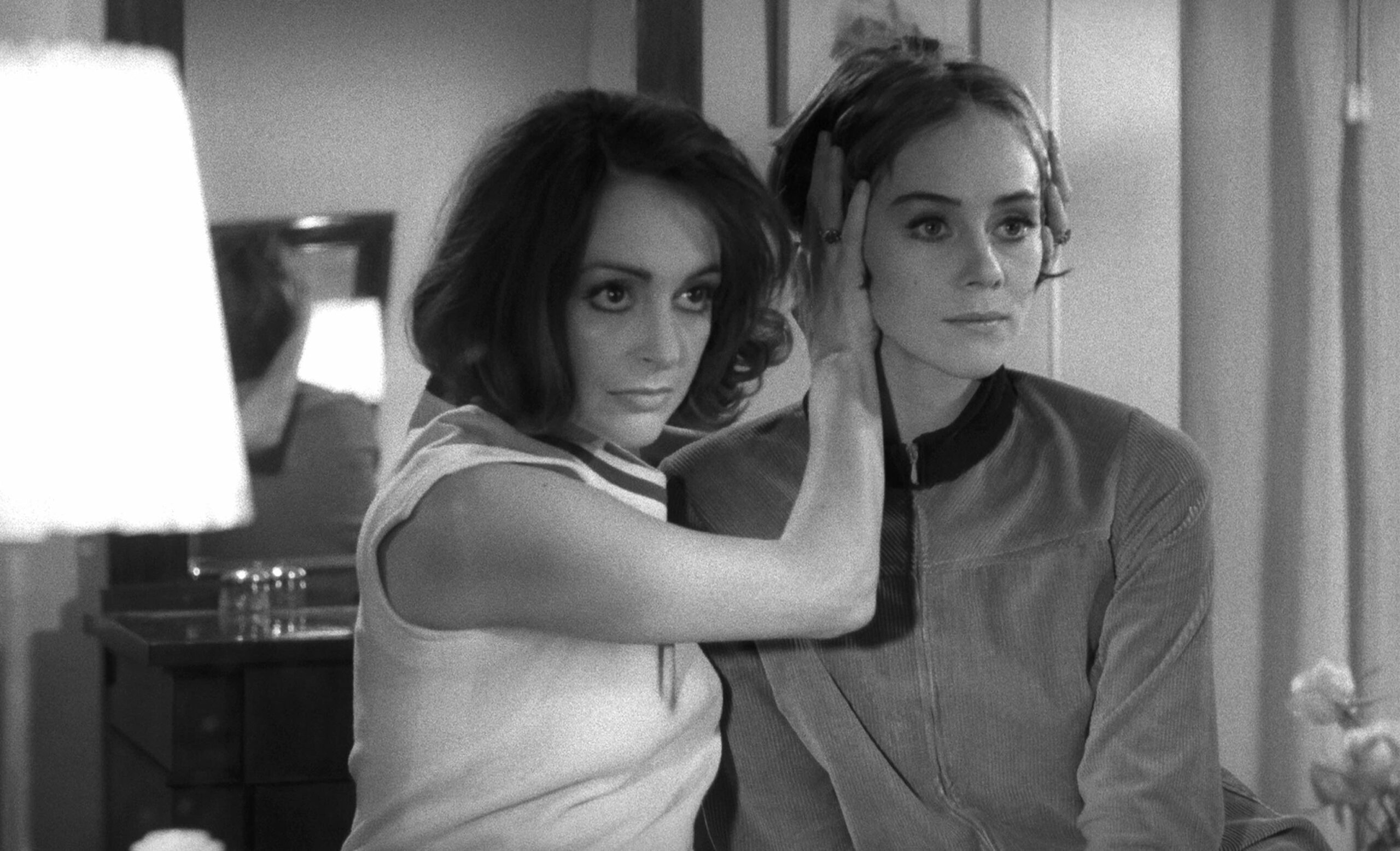 Two women sit in a bedroom, one pressing her hand against the side of the other's head, both looking off-screen
