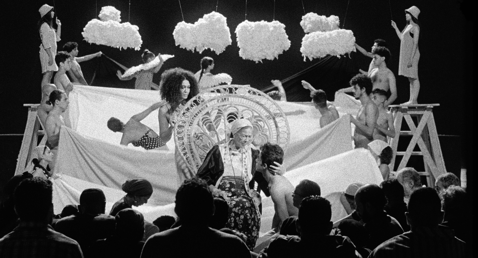 On a play stage an old woman holds a young man's head against a backdrop of ornate activity, including undulating white sheets to look like waves.
