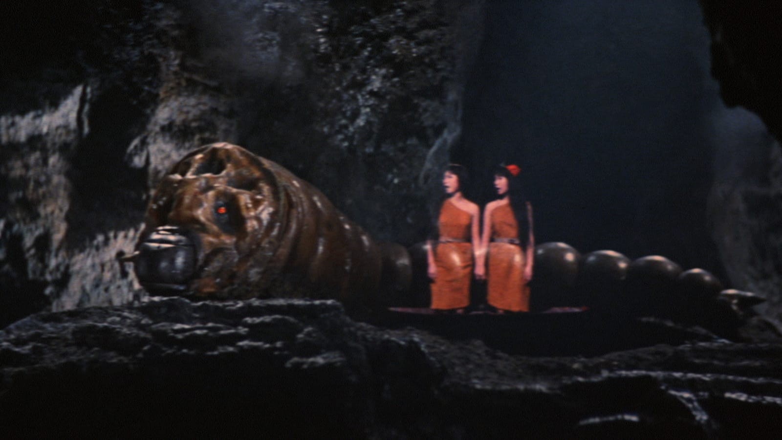 Two women in red joining hands stand in front of a giant worm beast.