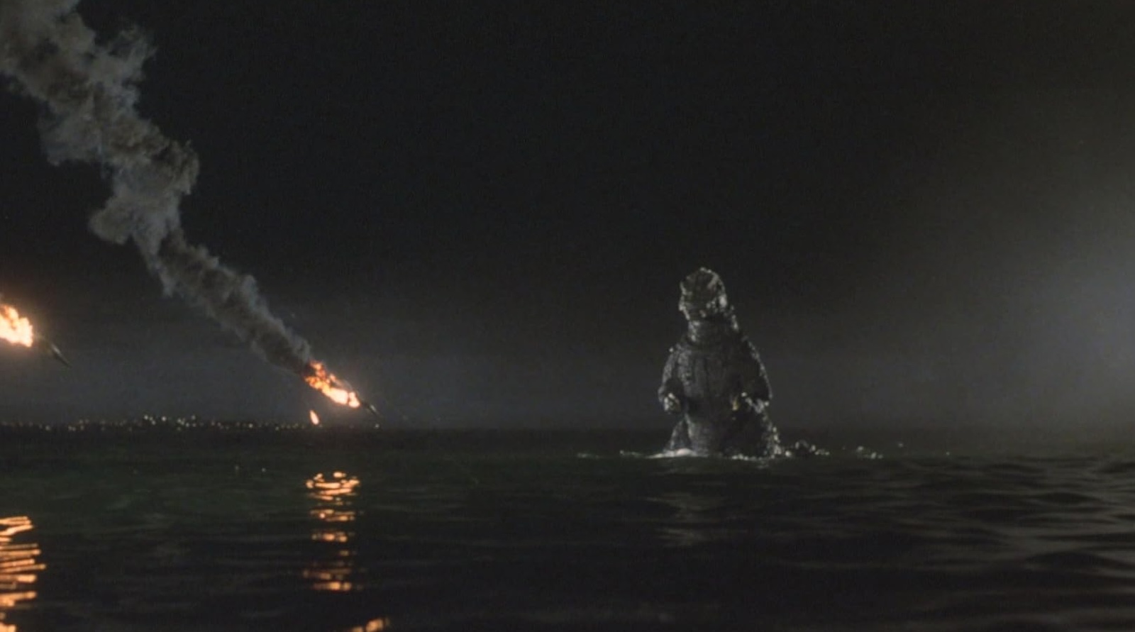 A giant rubber beast stands waist-high in water as a fiery missile sails past his head.
