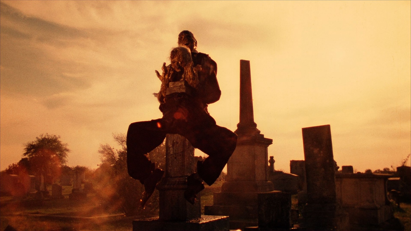 A corpse is arranged on a gravestone in a graveyard embracing another corpse against a yellow sky in silhouette.