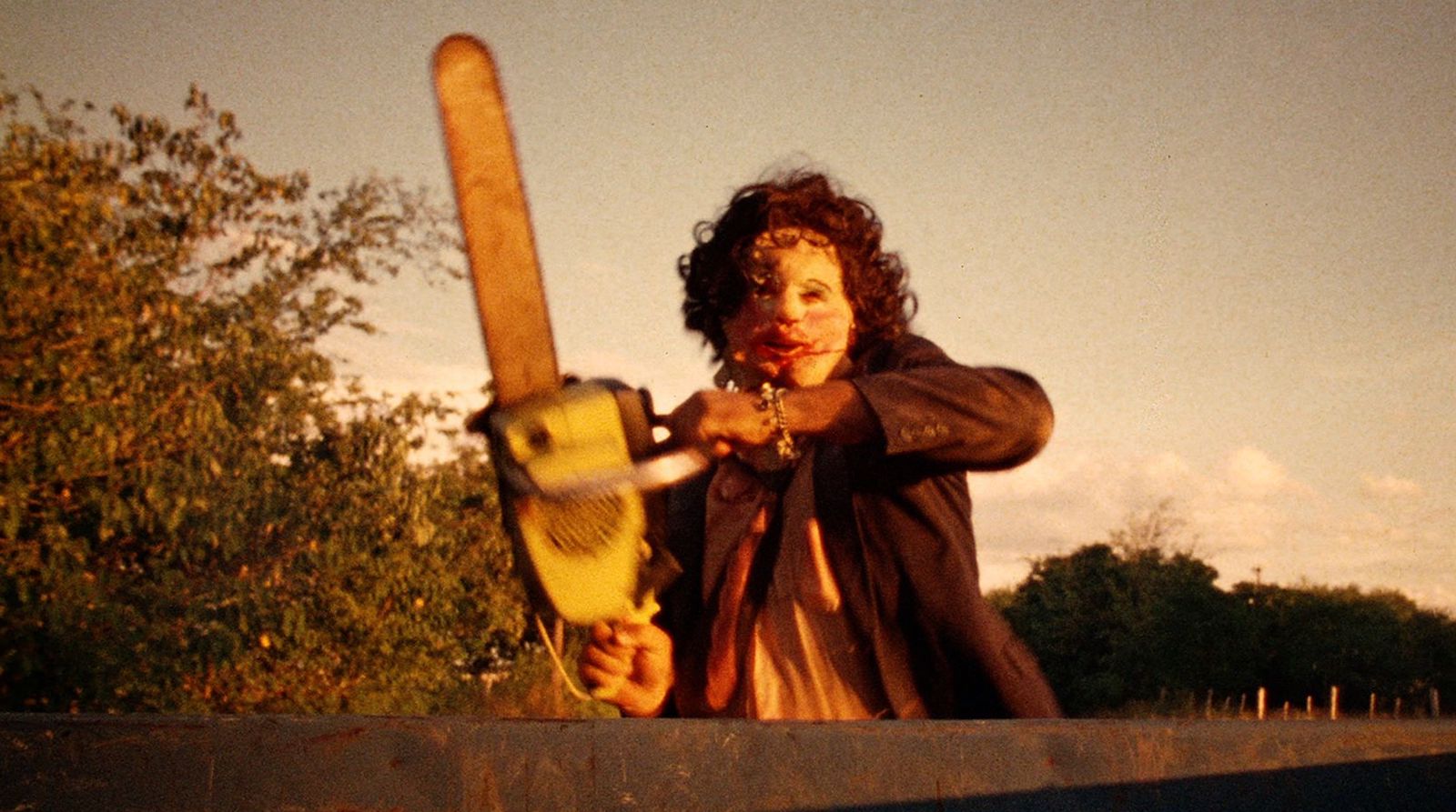 A deranged man wielding a chain saw and wearing a leather mask approaches the camera against a morning sky.