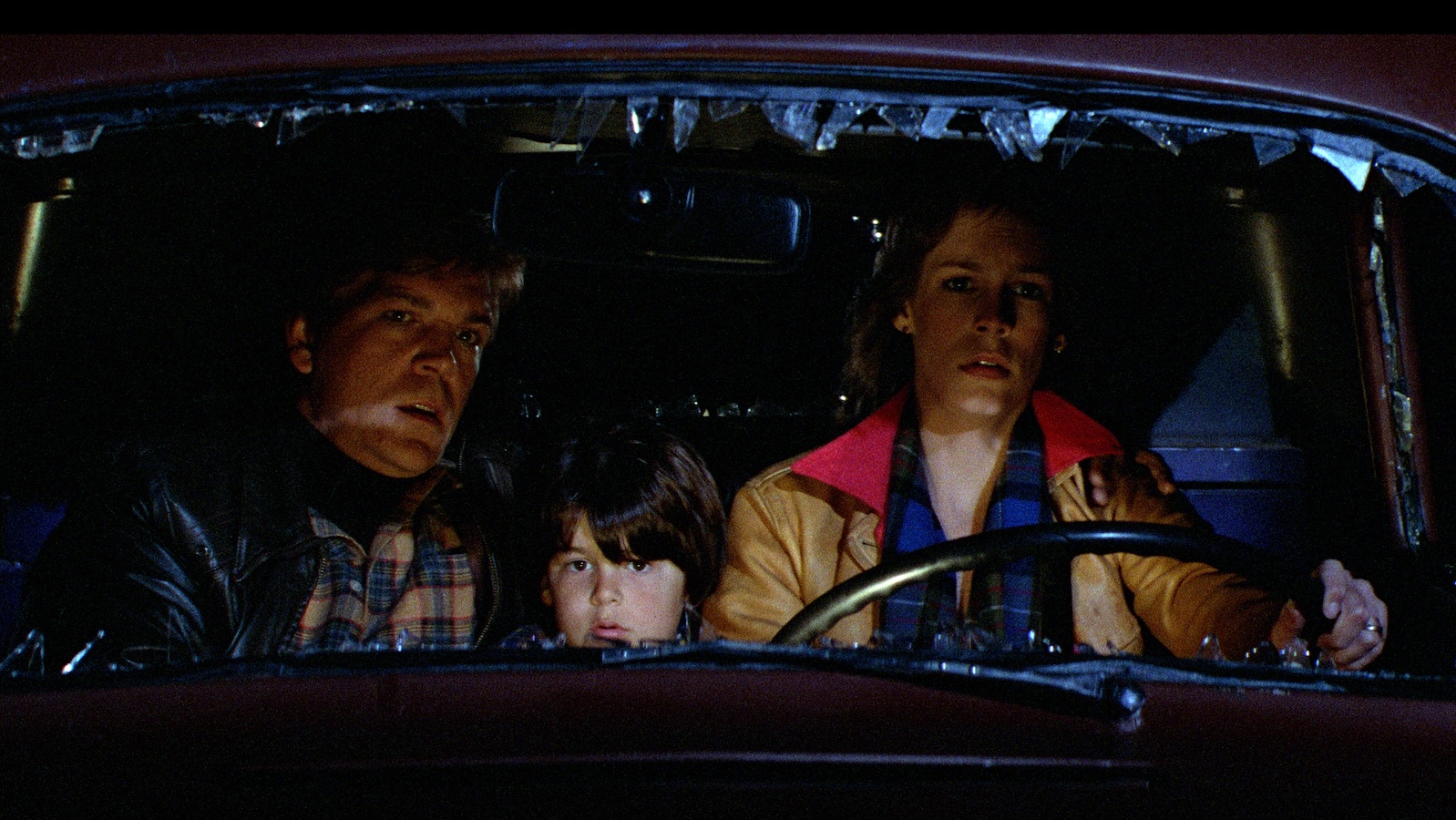 A man, woman, and child in a car at night with the windshield shattered and missing.