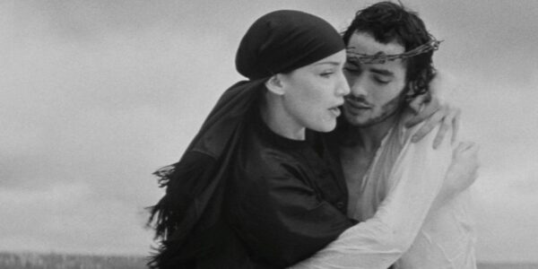 A woman in kerchief and man with crown of thorns embrace against a grim gray landscape.