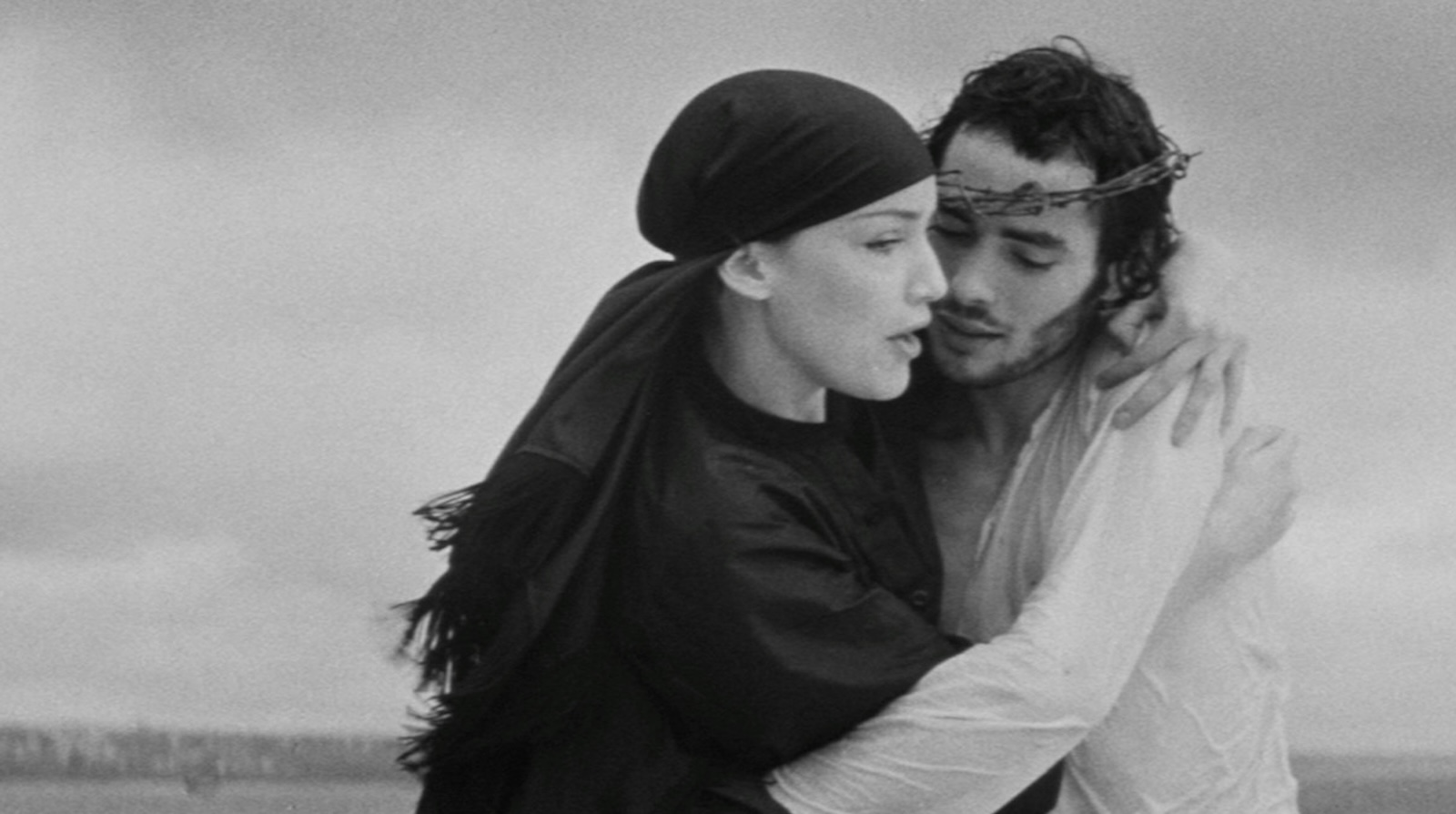 A woman in kerchief and man with crown of thorns embrace against a grim gray landscape.