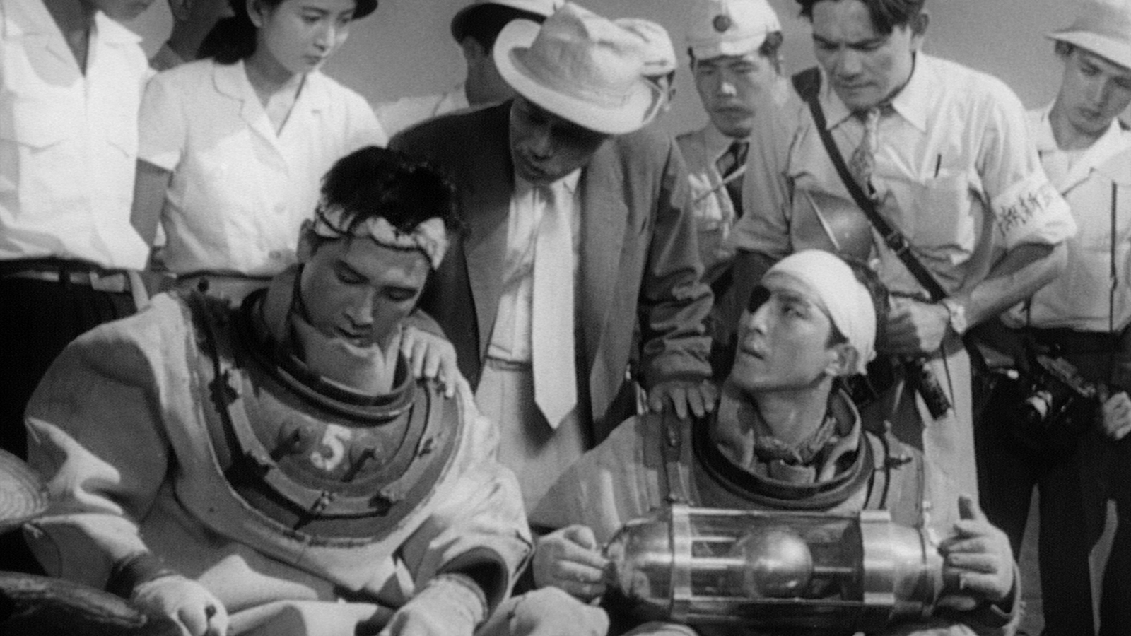 Two men wearing scuba diving gear sitting in front of standing people.
