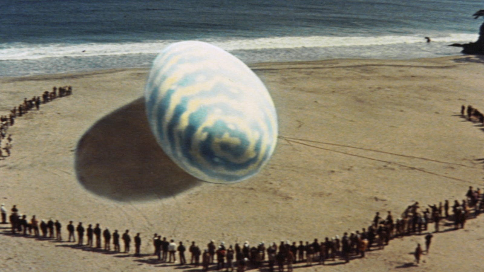 A gigantic blue and yellow egg on a beach surrounded by lines of people