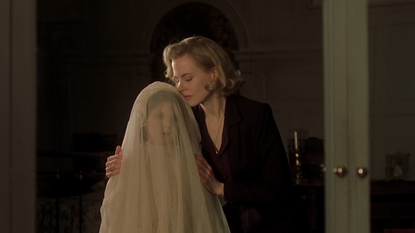 In a dark room, a blonde woman dressed in black embraces a young girl wearing a bridal veil that looks like a funeral shroud.