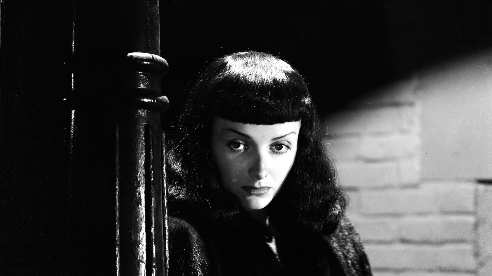 A sinister woman in black wigs with prominent bangs and fur coat stands in a dark alley at night against a street lamp