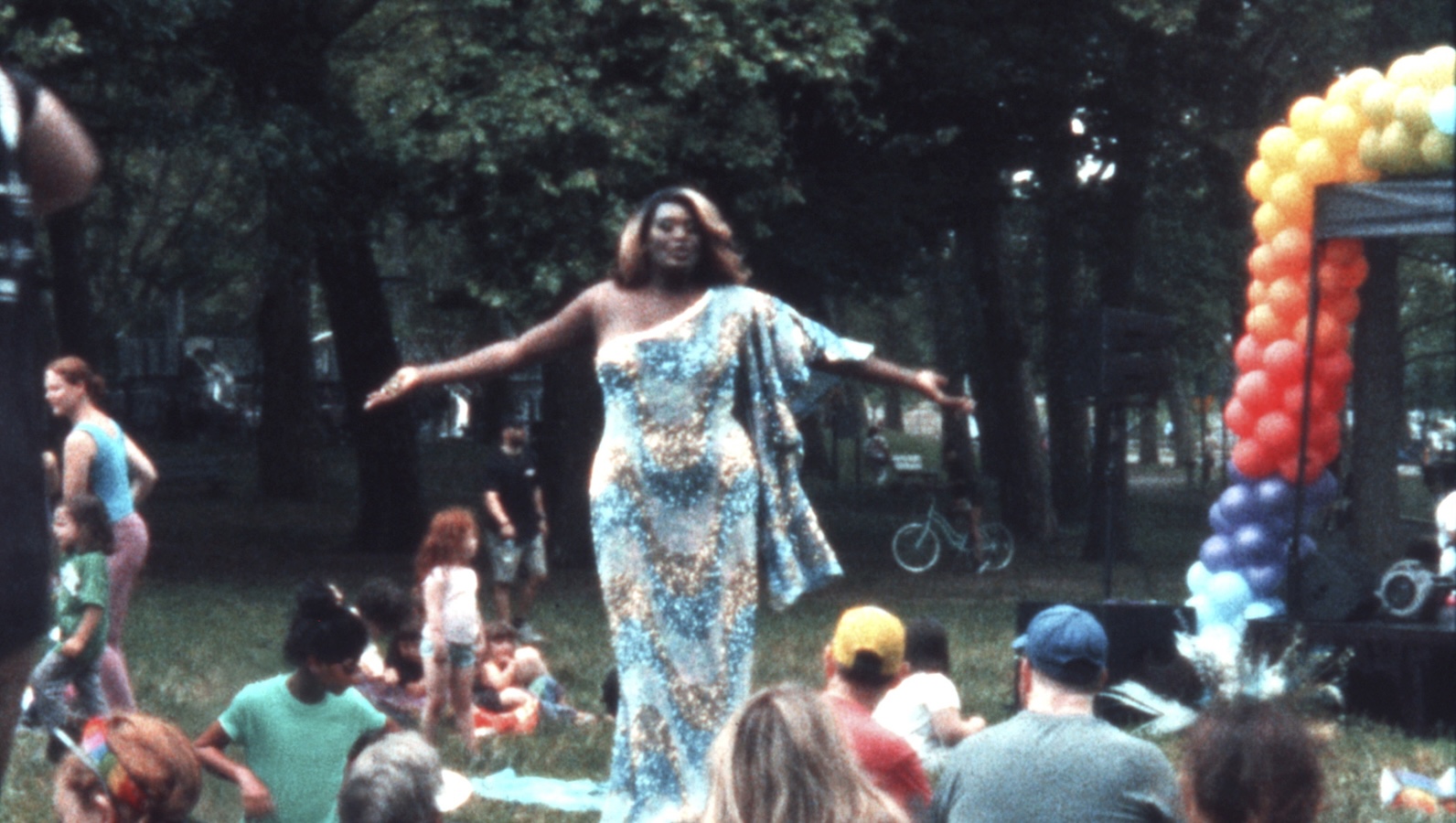 A dark-skinned drag performer in a blue dress stands in a park with arms outstretched, singing for people sitting on grass, next to rainbow colored balloons.