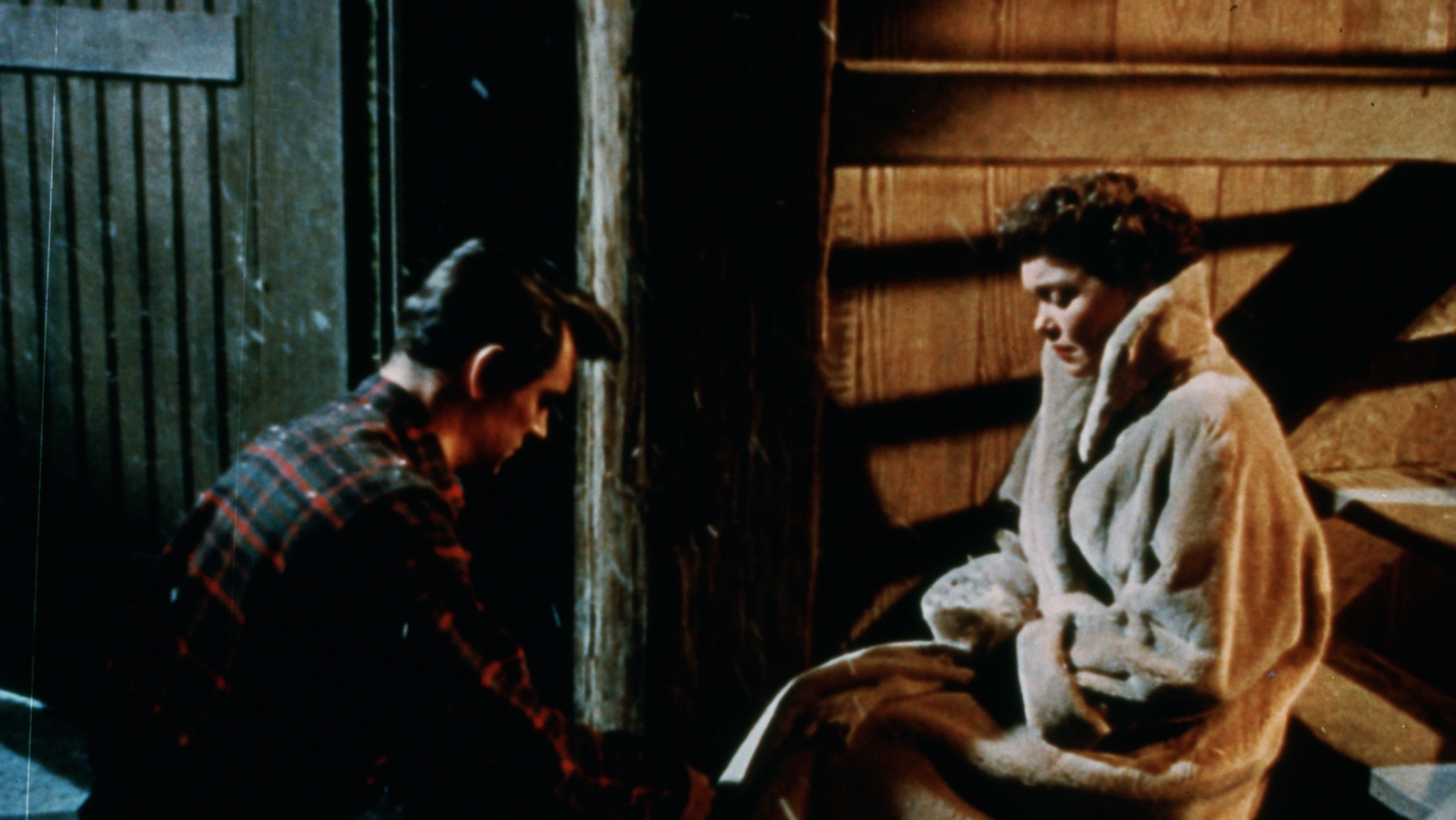 Near an open door with light snowflakes blowing, a man in red and green flannel crouches in front of a woman in a fur coat sitting on stairs. Both have serious, sad expressions.
