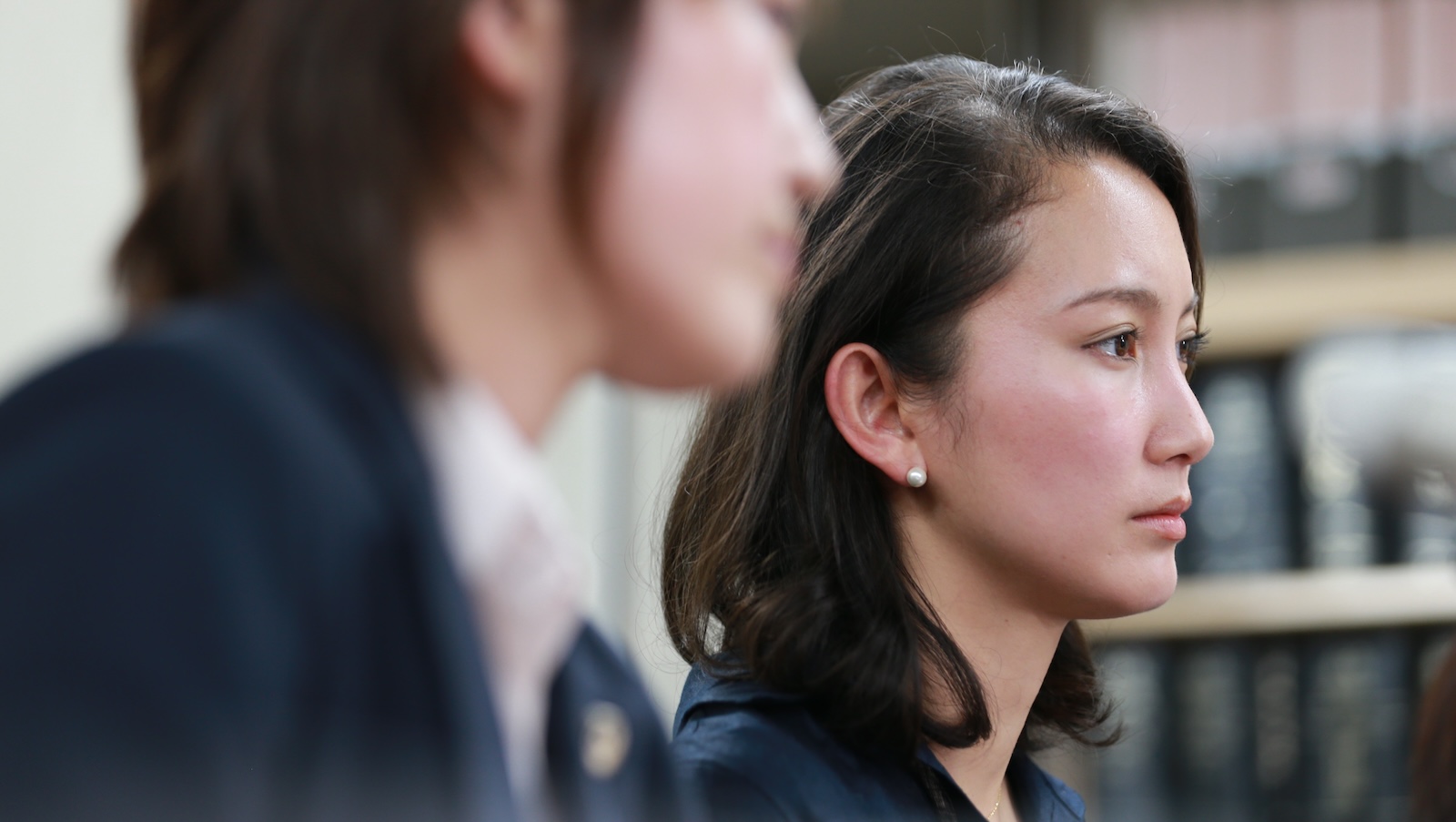 A woman in profile with dark long hair pulled behind her ear and a white pearl earring looks off with a serious expression, another woman out of focus in the foreground