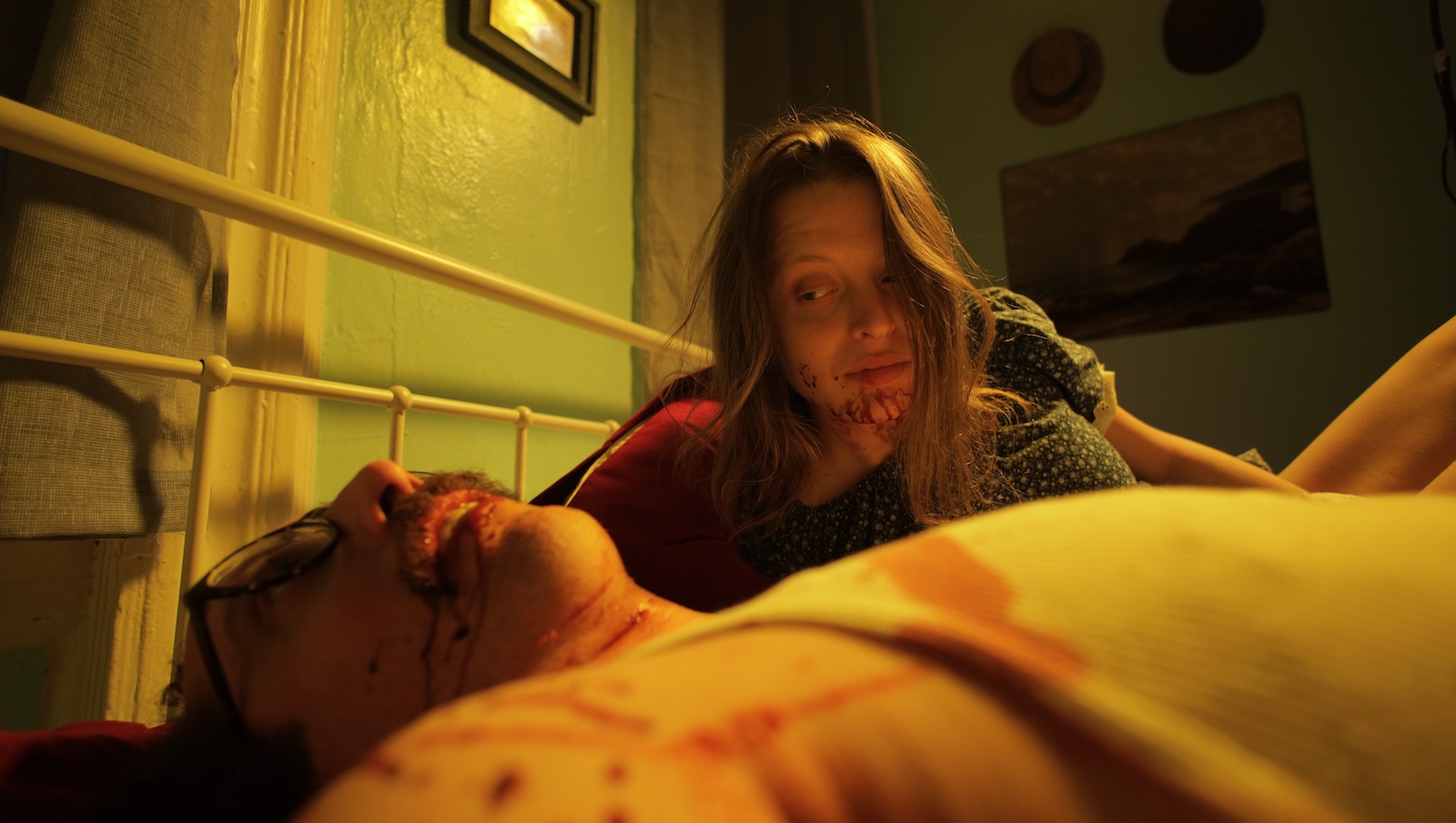 A blonde woman with a bloody mouth looks down smiling at a bloodied man in glasses lying on a bed in a soiled t-shirt